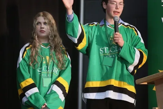 Two St. Clair College students (one being Samuel Smusiak) in green hockey jerseys on stage, one waving to the crowd while speaking into a microphone.